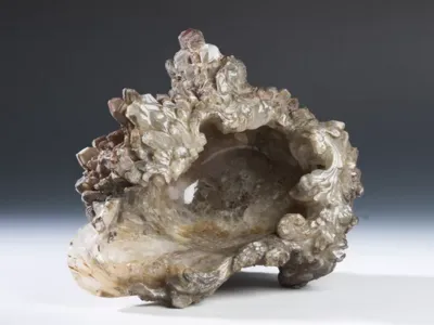 quartz ouvré dont l'intérieur lisse et poli contraste avec une paroi extérieure hérissée de cristaux bruts et de volutes baroques sculptées. La pierre présente des teintes allant du gris fumé au blanc laiteux, avec des reflets brillants. L'ensemble repose sur une surface blanche contre un fond dégradé gris foncé